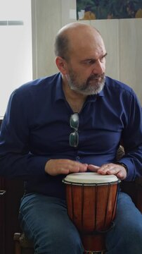 A focused and artistic mood is captured as a bald man with a beard intensely plays a djembe drum, his hands poised over the drumhead. This shot is ideal for depicting musical performance