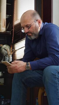 A contemplative or concerned mood is captured as a bald man with a beard and glasses sits, holding his smartphone with one hand and resting the other on his chin, suggesting a thoughtful