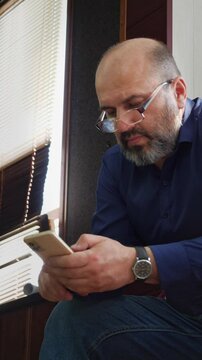 A focused and modern mood is captured as a bald man with a beard and glasses sits, intently looking down at his smartphone. This shot is ideal for depicting digital engagement, online communication