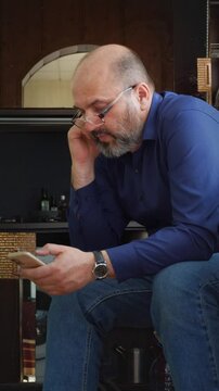 A focused and modern mood is captured as a bald man with a beard and glasses sits, intently looking down at his smartphone. This shot is ideal for depicting digital engagement, online communication