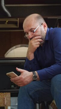 A focused and modern mood is captured as a bald man with a beard and glasses sits, intently looking down at his smartphone. This shot is ideal for depicting digital engagement, online communication