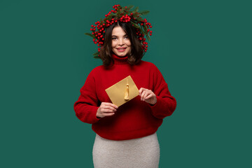 Cheerful woman in a red sweater holding a golden envelope, wearing a berry wreath, smiling