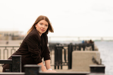 Smiling woman in brown blazer looking at camera near waterfront. Natural feel with soft light. Casual, confident pose and expression add genuine and approachable vibe