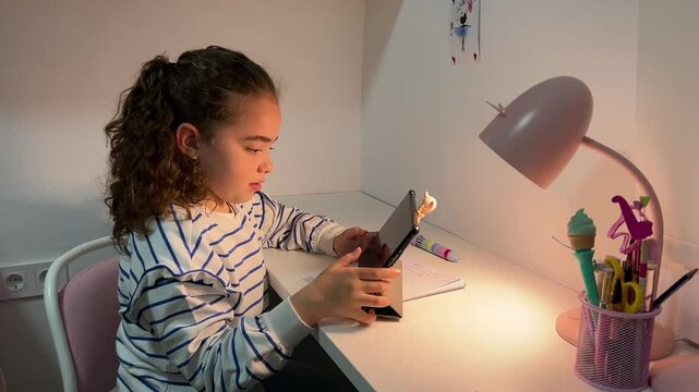 Little girl using a tablet at her study desk, exploring digital content with curiosity and focus, highlighting early childhood interaction with technology in a cozy home environment.