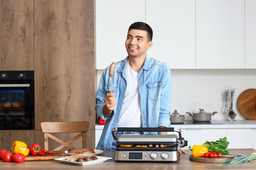 Young man with cooked tasty sausages and vegetables on modern electric grill at table in kitchen