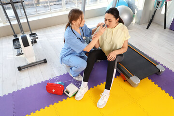 Female doctor with inhaler giving young woman first aid in gym