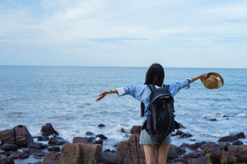 Young Asian woman enjoying the ocean breeze while standing on rocky shore with arms outstretched...