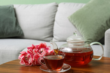 Transparent teapot and cup of hot tea with pink tulip flowers on table in living room