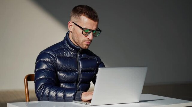Focused man working on laptop cold balcony wearing down jacket