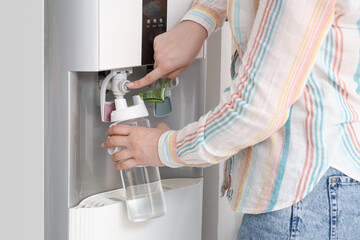 Woman filling bottle with filtered water from cooler near white wall