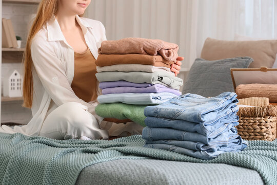 Young happy woman with stacks of stylish clothes and wicker basket sitting on bed at home, closeup