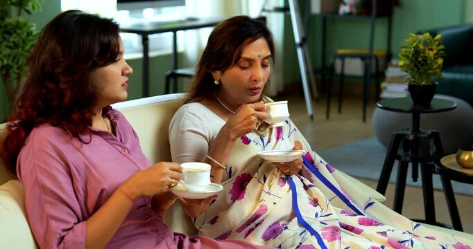 Senior Mother Daughter drinking tea, Indian asian old mother in saree with young daughter sitting together on sofa in modern living room enjoying hot morning beverage while chatting bonding joyfully