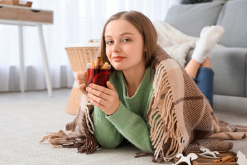 Beautiful young woman in warm sweater with glass cup of hot mulled wine and blanket lying on floor...