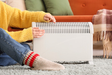 Young woman sitting on floor near modern convector heater at home