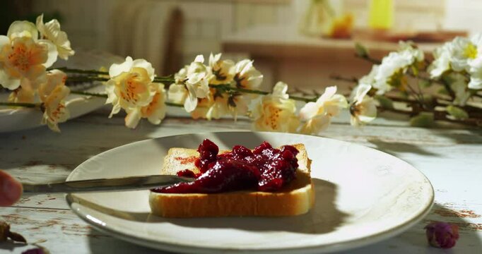 hand uses a butter knife to spread rich berry strawberry jam over a slice of toast on a white ceramic plate, set on a weathered wooden table adorned with delicate cream blossoms in warm morning light.