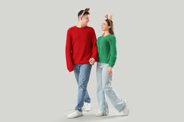 Cute young couple in warm sweaters and Christmas headbands walking on grey background