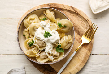 Bowl of delicious boiled dumplings with parsley and sour cream on white wooden background