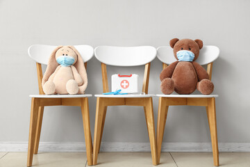 Cute toys in medical masks with first aid kit and stethoscope sitting on chairs near grey wall