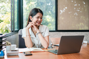 A young woman smiling as she works on her laptop in a bright, modern office with large windows and lush greenery outside.