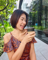 Young cheerful Asian woman in a striking orange-patterned blue dress holds a paper cup of coffee, embracing an eco-conscious urban lifestyle