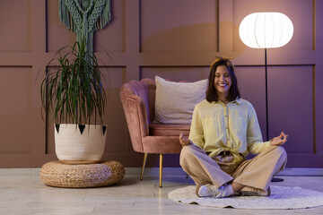 Young woman meditating near pink armchair at home in evening