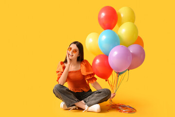 Beautiful teenage girl in sunglasses with colorful air balloons sitting on yellow background