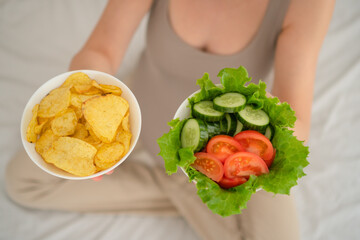 Pregnant woman comparing fresh vegetable salad with potato chips. Realistic moment of food choices...