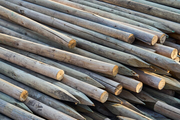 A stack of wooden stakes with sharpened ends, arranged in a pile at a construction or agricultural site. The natural texture and grain of the timber are clearly visible, with warm sunlight casting sof