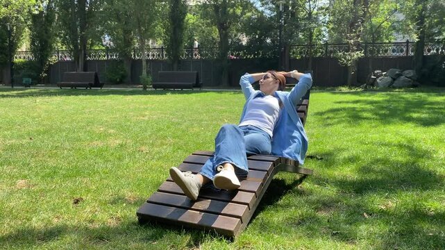 A person fully relaxed, reclining on a wooden lounge chair on a vibrant green lawn, with their hands behind their head and wearing sunglasses. The expansive, sun-drenched park, bordered by trees