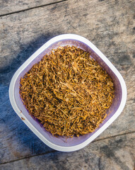A plastic container filled with dried shredded tobacco sits on a rustic wooden surface, ready for...