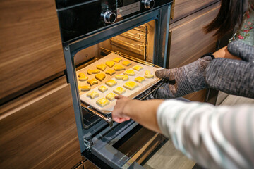 Close up of unrecognizable mom hands taking baking pan of golden cookies out of oven in kitchen while daughter pointing to biscuits with finger
