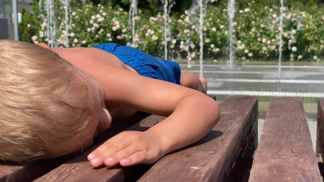 A young child with fair hair lies face down, seemingly resting, on a wooden park bench, with a backdrop of lively water fountains and blooming rose bushes in a sunny Simferopol park.