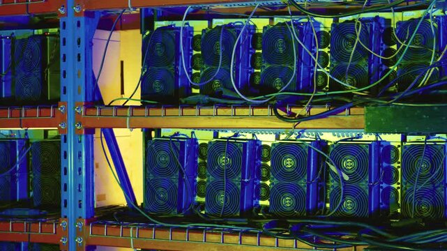 Multiple ASIC bitcoin miners with prominent cooling fans and tangled wiring mounted on metal shelves in a server rack. Close up pan right