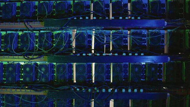 Horizontal shelves of ASIC bitcoin mining units with power and data cables, blue lighting. Tilt-down view reveals multiple stacked rows