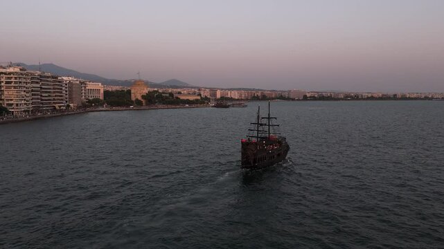 Rear view of pirate ship sailing in Thessaloniki with with white tower at background during sunset, drone shot.