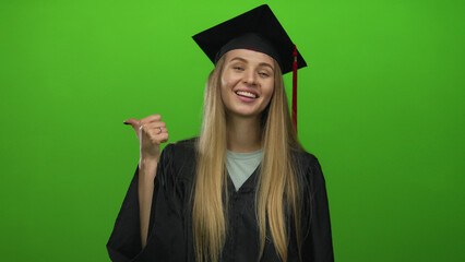 Young caucasian woman in graduation gown smiles against an isolated green wall background,...