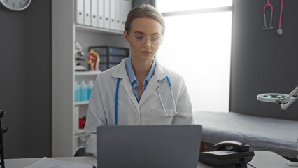 Female doctor working on laptop in clinical office setting with medical equipment, wearing glasses...