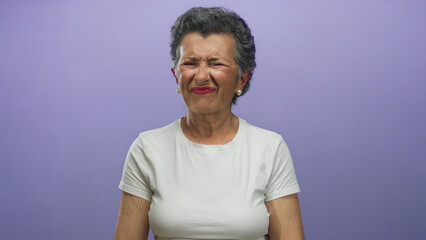 Senior woman with grey hair in white shirt grimacing against a purple background, capturing a...