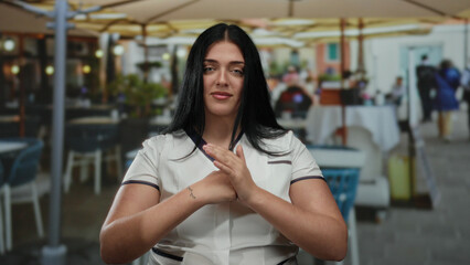 Young woman with long black hair sitting on an outdoor restaurant terrace, gesturing with her...