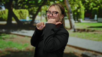 Fototapeta premium Woman in glasses smiling with thumbs pointing backwards in a park setting.