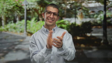 Caucasian man clapping hands joyfully in a park setting with greenery and dappled sunlight, wearing...