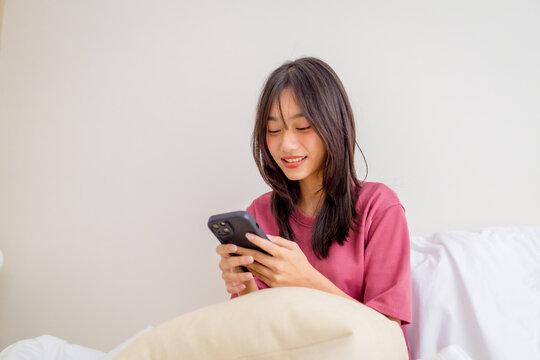 Asian young girl using smartphone texting and chatting with friend in bedroom at home. Relaxing woman wearing pink shirt sit on the bed using mobile phone to communicate in the morning at house.