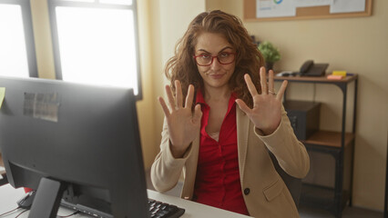 Woman gesturing with hands in office setting with computer, wearing glasses and red outfit,...