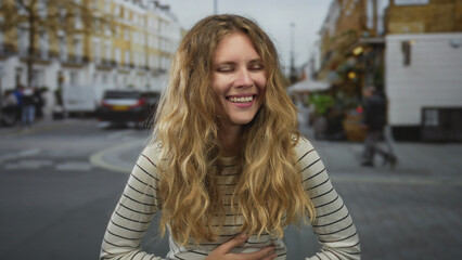 Fototapeta premium Woman raises hand while laughing on a street lined with buildings as blonde young subject smiling and clutching stomach in a joyful casual moment.