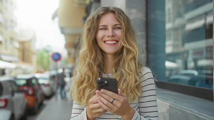 Vibrant portrait captures a woman holding a phone with a smiling expression on a city street, her blonde hair and young face illuminated outdoors as she checks a smartphone over a striped shirt.
