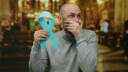 Man holding cancer awareness ribbon in church setting, exhibiting emotional response with...