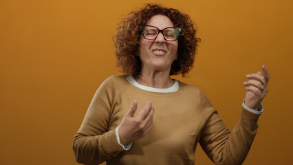 Middle-aged hispanic woman playfully mimics playing guitar against an isolated orange wall...