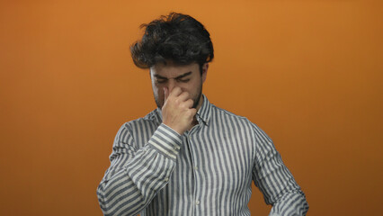 Young hispanic man with a beard covering his nose from a bad smell against an orange background,...