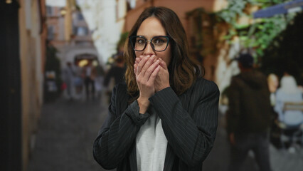 Fototapeta premium Hispanic woman in glasses on a bustling city street covers mouth in surprise, surrounded by blurred people and urban environment, creating a vivid city life scene.