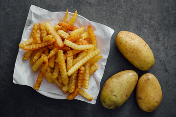 Crispy Crinkle Cut French Fries with Fresh Raw Potatoes on Dark Background